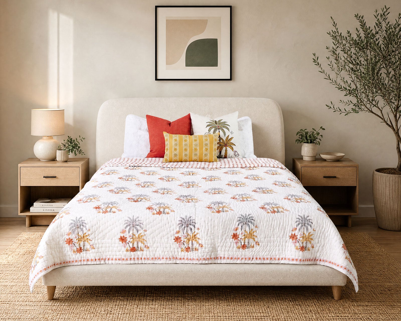 A styled bedroom scene showing a bed dressed in a white quilted coverlet with a repeating palm tree print in muted orange, gray, and green. The bed is styled with white, red, and yellow accent pillows, flanked by matching wooden nightstands with lamps and a potted tree against a warm beige wall.