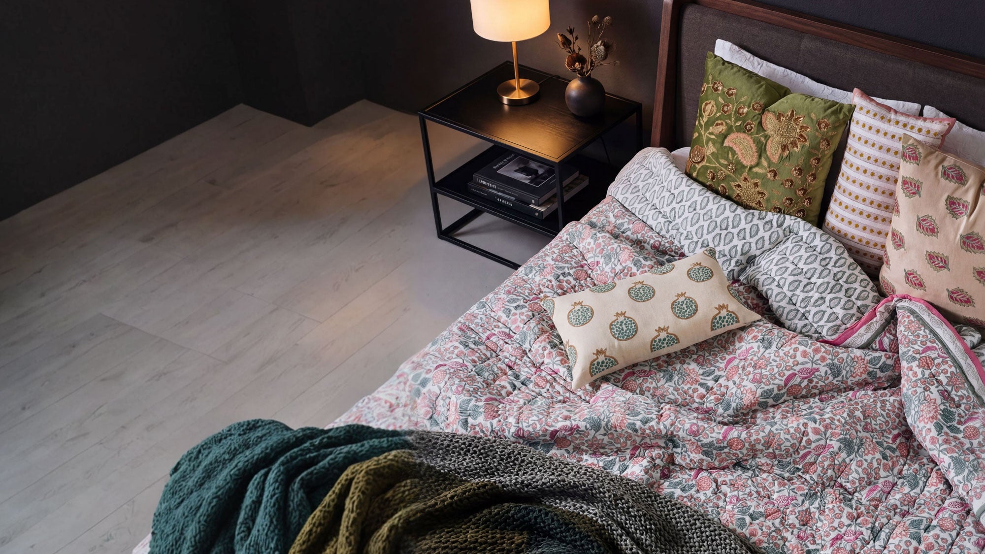 Bedroom with floral bedding, patterned pillows, and a side table with a lamp.